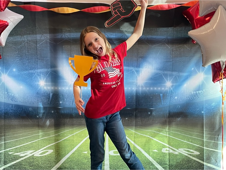 fourth grader holds gold paper trophy while standing in front of a football field tapestry