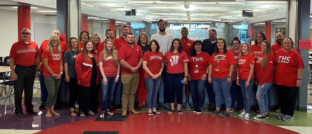 seventh and eighth grade TMS staff pose for a quick pic  in front of glass display cases. they all wear red shirts