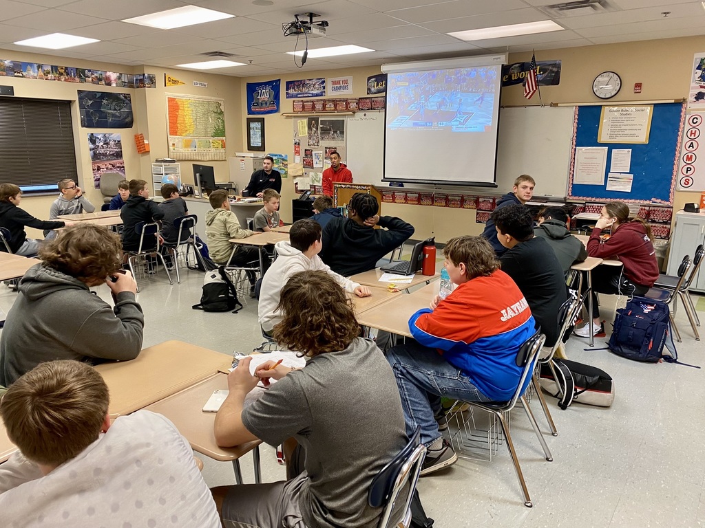 classroom of middle school students sitting at desks. a projector screen playing basketball highlights hangs from the ceiling at the front of the room