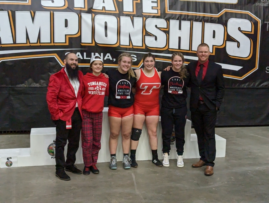 Four girls wrestling team members are flanked on each side by a coach, standing in front of state championships banner