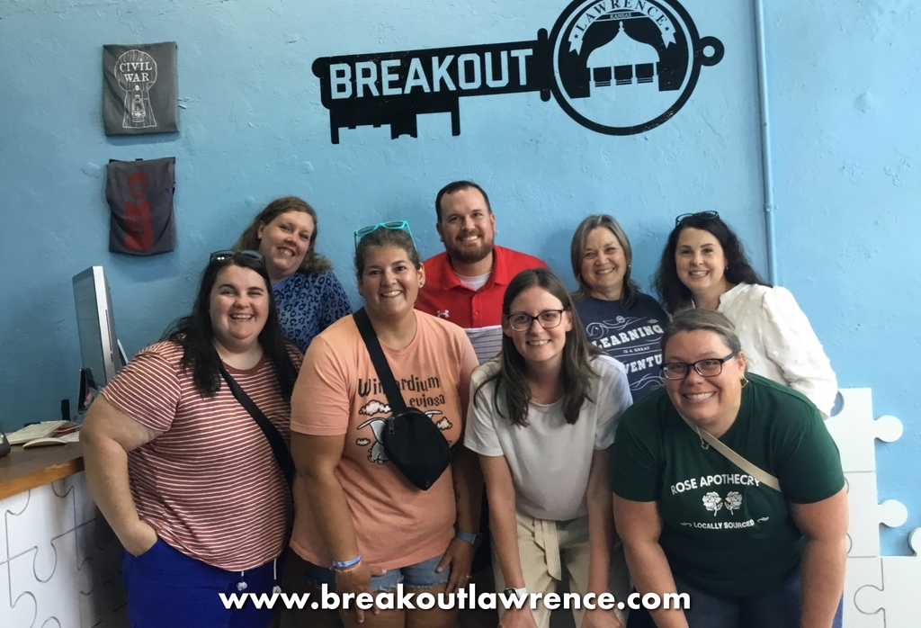 eight adults stand in front of a blue concrete wall with a black key graphic that says Breakout Lawrence