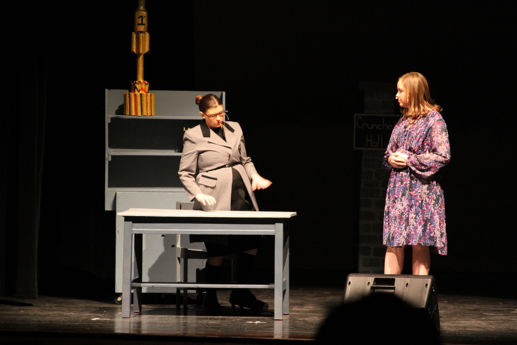 Two middle school girls stand on stage. One wears a black and gray suit; the other a pink and purple flowered dress.