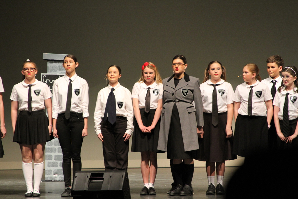 Eight middle school students wearing black skirts or pants and white shirts with black ties sing on stage as a student in a black and gray suit stands in front of them.