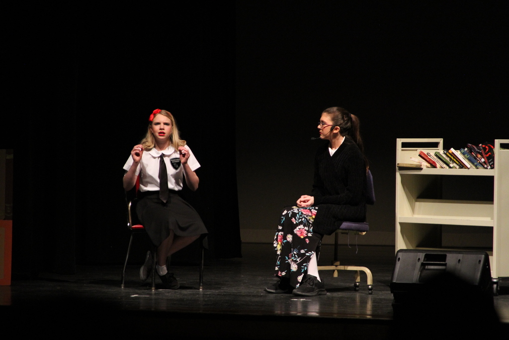 Two students sit on chairs on stage as they perform Matilda. A rolling cart of books sits nearby.
