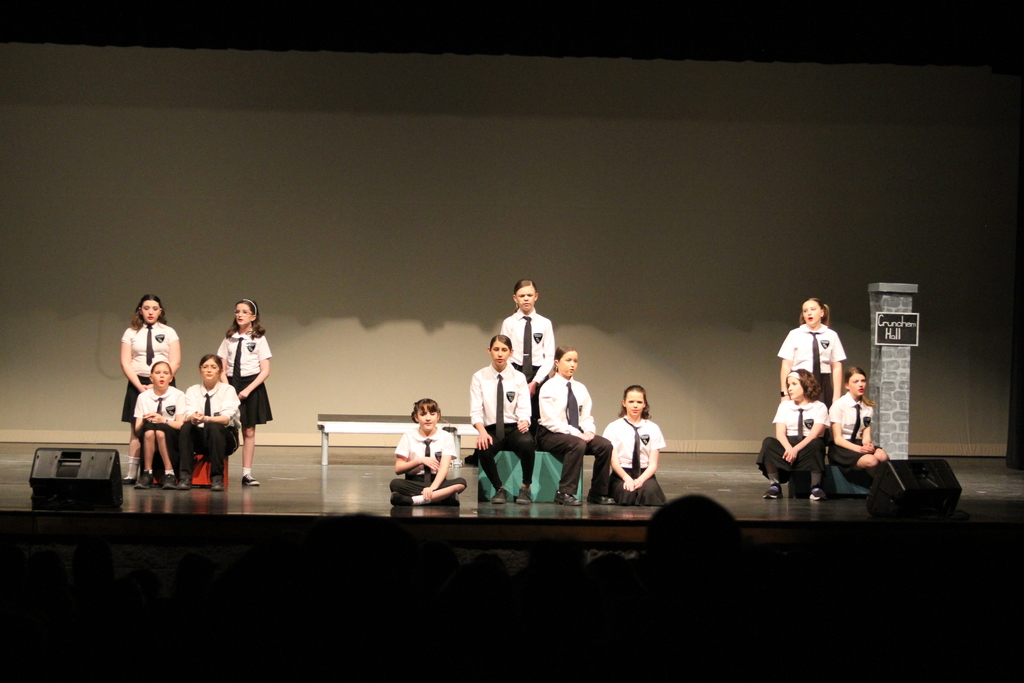 Twelve middle school students wearing black skirts or pants and white shirts with black ties sing on stage.