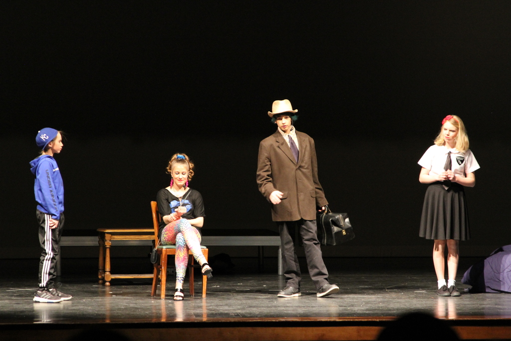 Four middle school students stand on stage during Matilda. One wears a brown suit coat, another a black shirt and leggings, another black track pants and blue hoodie, and the last a black skirt and white shirt with black tie.