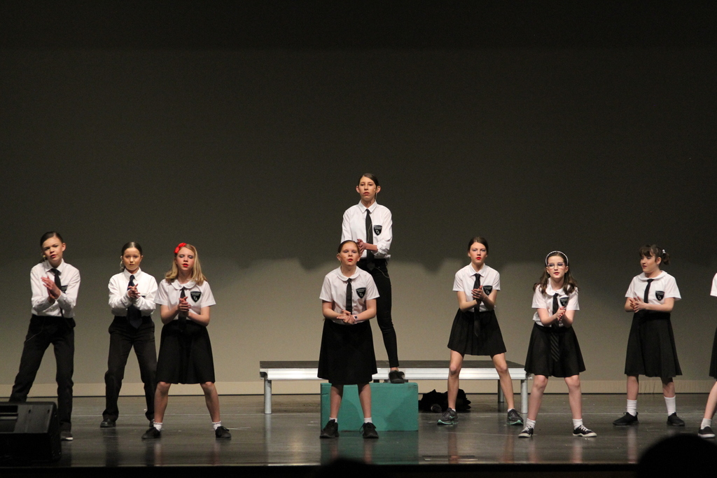 Eight middle school students wearing black skirts or pants and white shirts with black ties sing on stage.