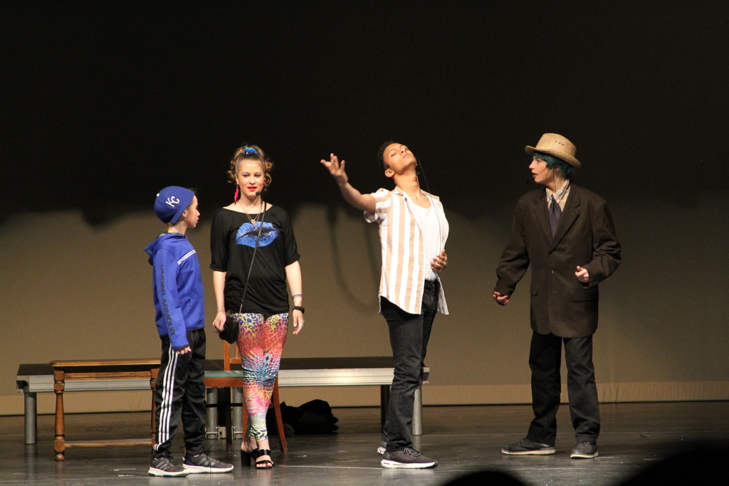 Four middle school students stand on stage during the performance of Matilda The Musical JR. One in a white and orange striped shirt gestures with his right hand.