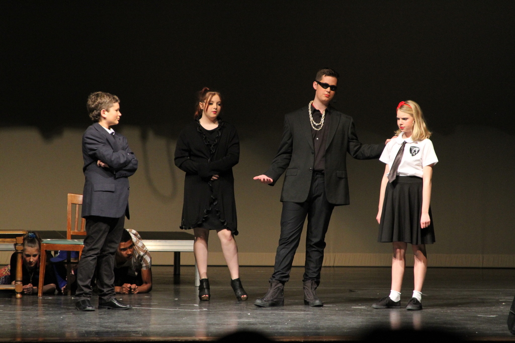Four middle school students stand on a stage during a performance. They all wear black. One wears sunglasses.