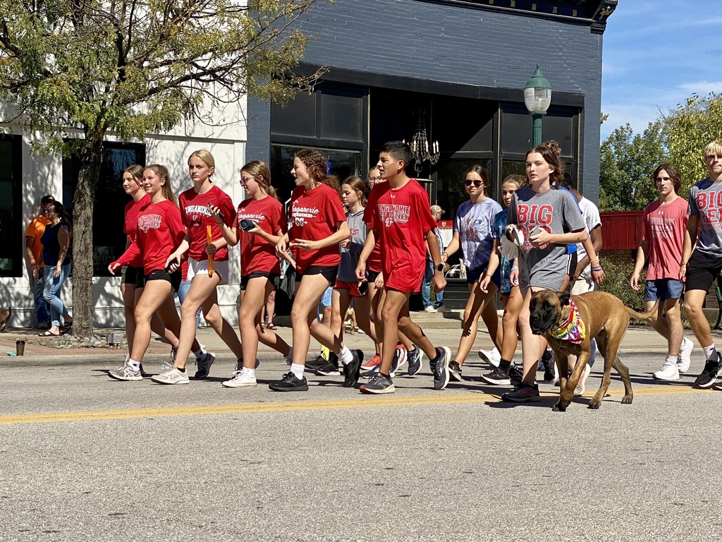 cross country members wearing red shirts walk down the street during the parade